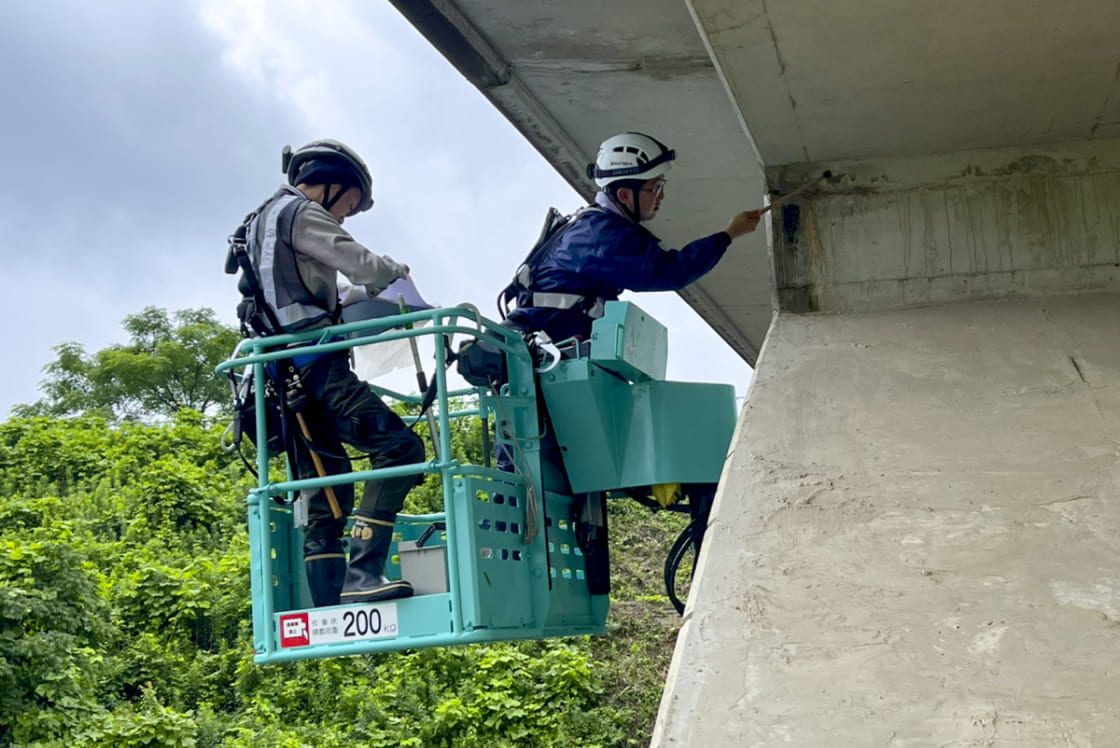 高所作業車の上で、橋の下部を点検・作業している2人の作業員。